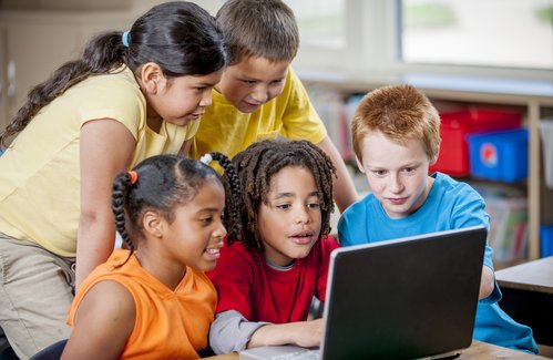 Das Bild zeigt eine Gruppe von fünf Kindern, die um einen Laptop versammelt sind. Sie schauen gemeinsam gespannt auf den Bildschirm, während sie offensichtlich etwas Interessantes betrachten oder lernen. Die Szene spielt sich in einem hellen Raum ab, vermutlich in einem Klassenzimmer oder einer Lernumgebung.