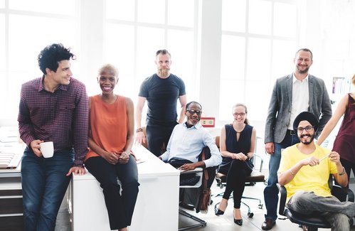 Eine Gruppe von zwölf Personen sitzt und steht in einem modernen Büro mit hellen, großen Fenstern. Sie wirken entspannt und lächelnd, während sie in die Kamera schauen. Einige halten Tassen in der Hand. Im Hintergrund sind Regale mit Ordnern und Büchern sowie Computerarbeitsplätze zu sehen. Die Szene vermittelt eine positive und kollegiale Arbeitsatmosphäre.