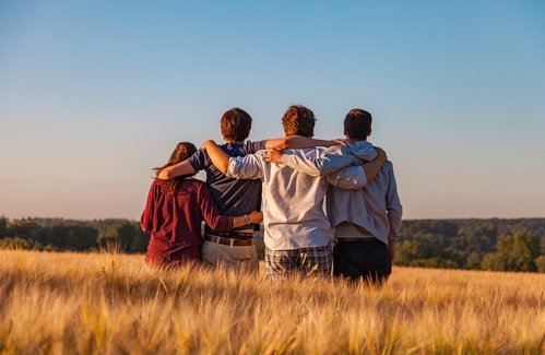 Vier junge Menschen stehen in einem Feld mit goldenem Getreide, Arm in Arm, und schauen in die Ferne. Die Sonne steht tief am Himmel und taucht die Szene in warmes Licht. Der Himmel ist klar und blau, und im Hintergrund sind Wälder zu sehen.