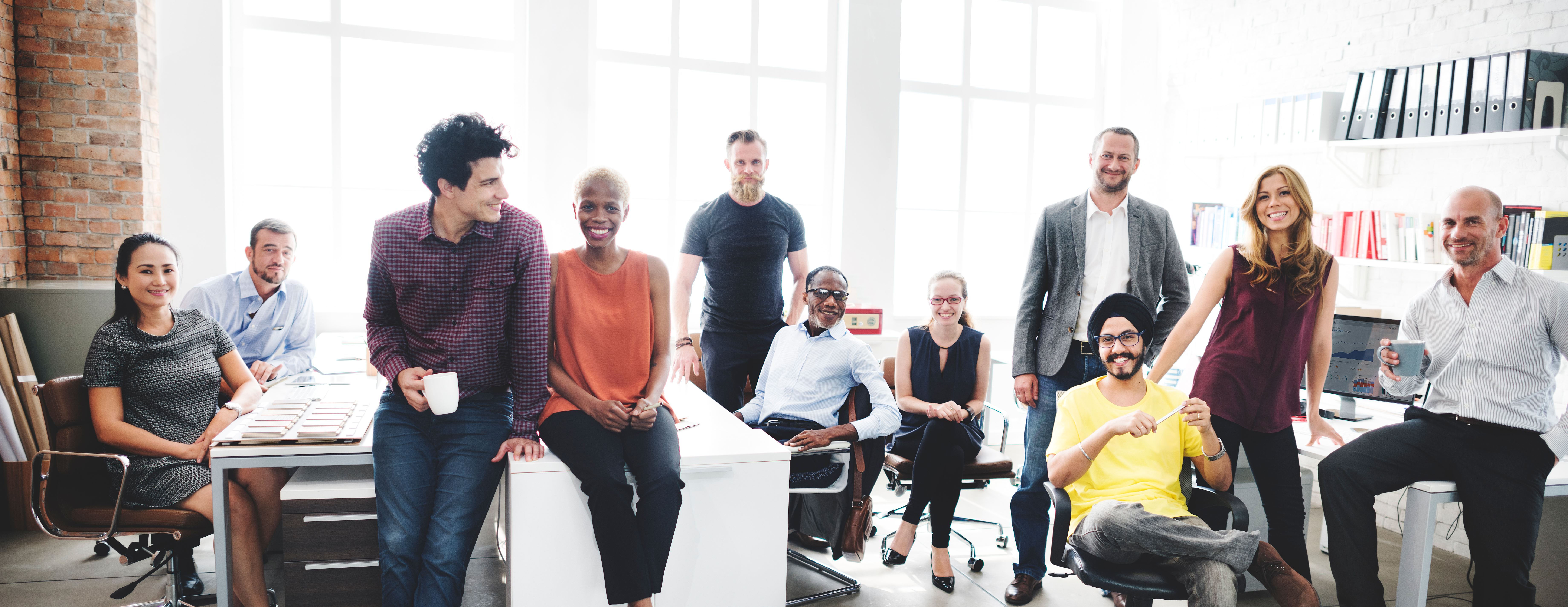 Eine Gruppe von zwölf Personen sitzt und steht in einem modernen Büro mit hellen, großen Fenstern. Sie wirken entspannt und lächelnd, während sie in die Kamera schauen. Einige halten Tassen in der Hand. Im Hintergrund sind Regale mit Ordnern und Büchern sowie Computerarbeitsplätze zu sehen. Die Szene vermittelt eine positive und kollegiale Arbeitsatmosphäre.